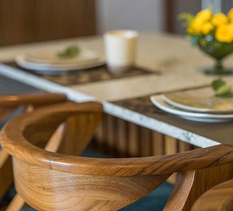 Curved teak dining chairs at a stone-topped table set with plates and yellow roses
