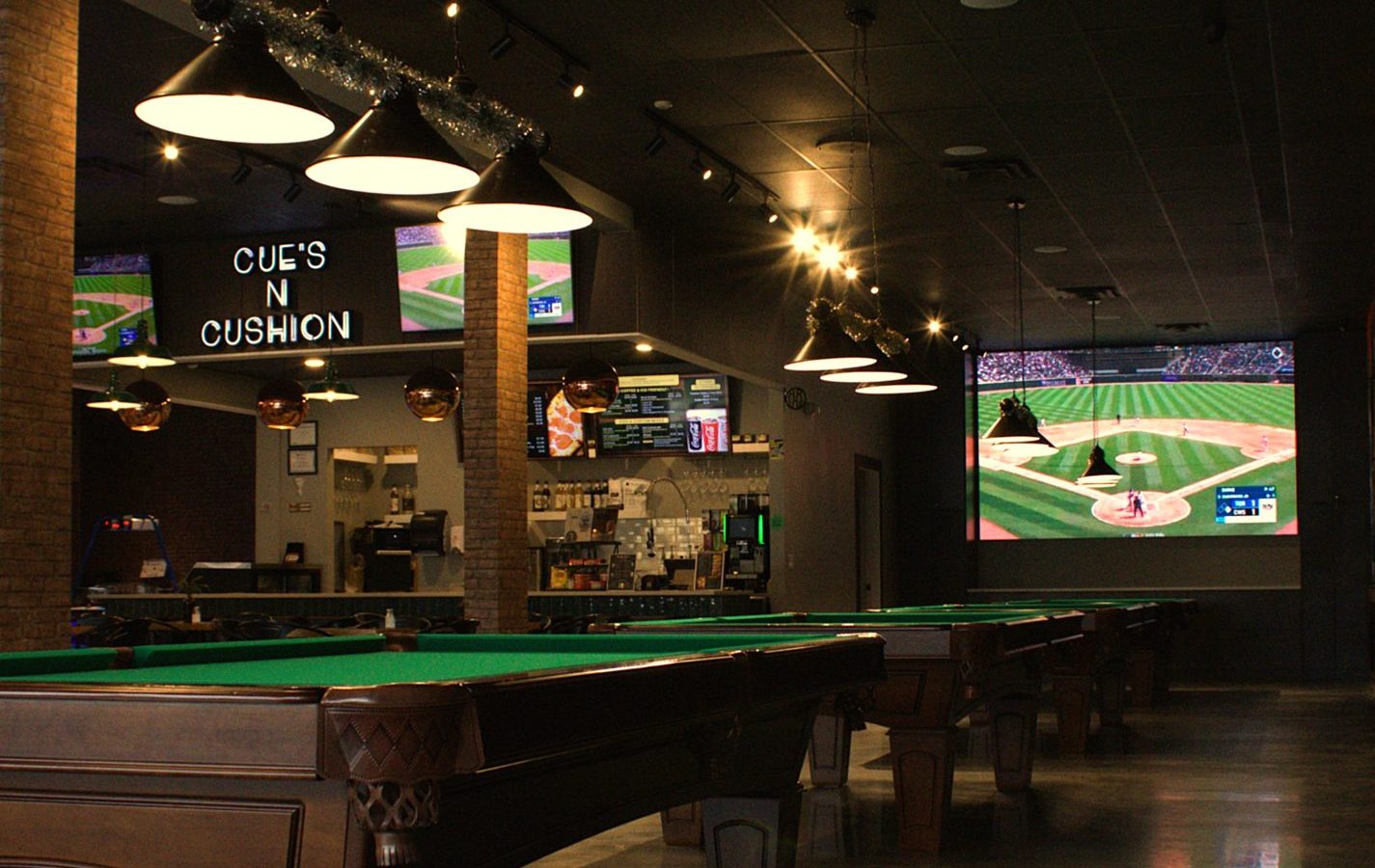 Pool tables under pendant lighting with brick columns at Cue's N Cushion