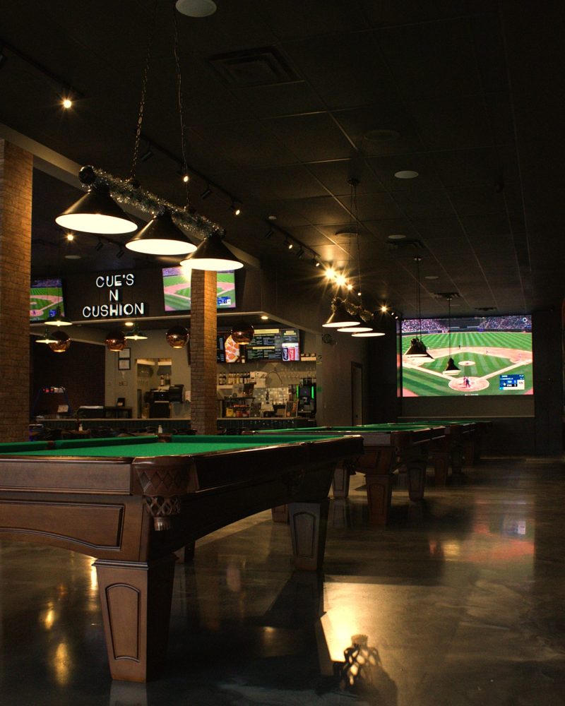 Pool tables under pendant lighting with brick columns at Cue's N Cushion